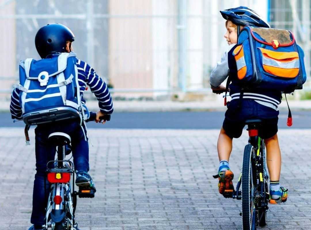 Two children wearing helmets ride bicycles on a paved path, carrying large backpacks, as they travel side by side in an urban setting.