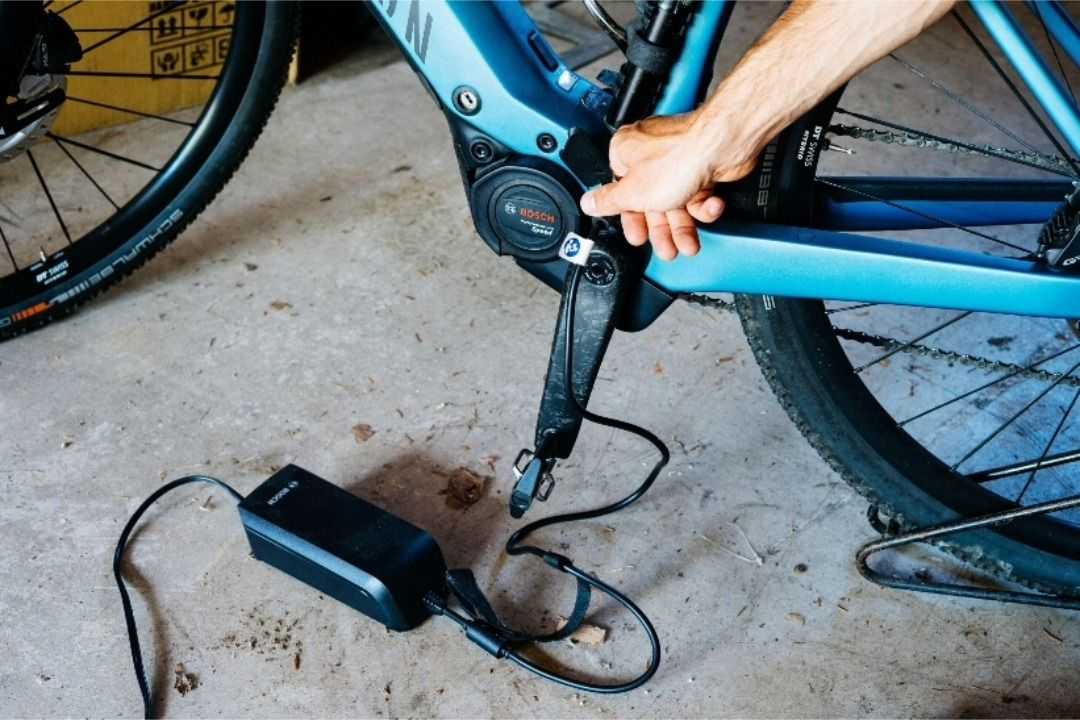 Close-up of an e-bike being plugged into a charger, with a hand connecting the charging cable to the bike’s battery system in a garage or workshop setting.