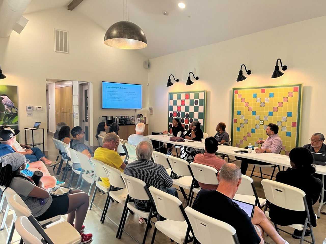 Community meeting in a bright room; attendees sit in rows facing a panel at tables, with a presentation screen and two large board displays on the wall behind the speakers.