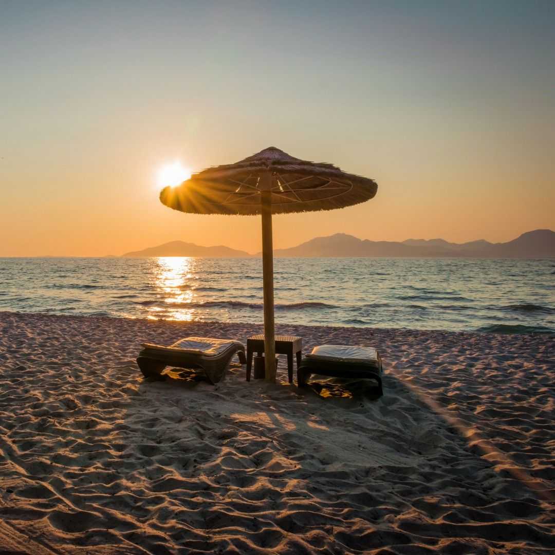 Beach scene at sunset with two lounge chairs and a straw umbrella facing calm ocean water, sunlight reflecting on waves and distant mountains on the horizon.